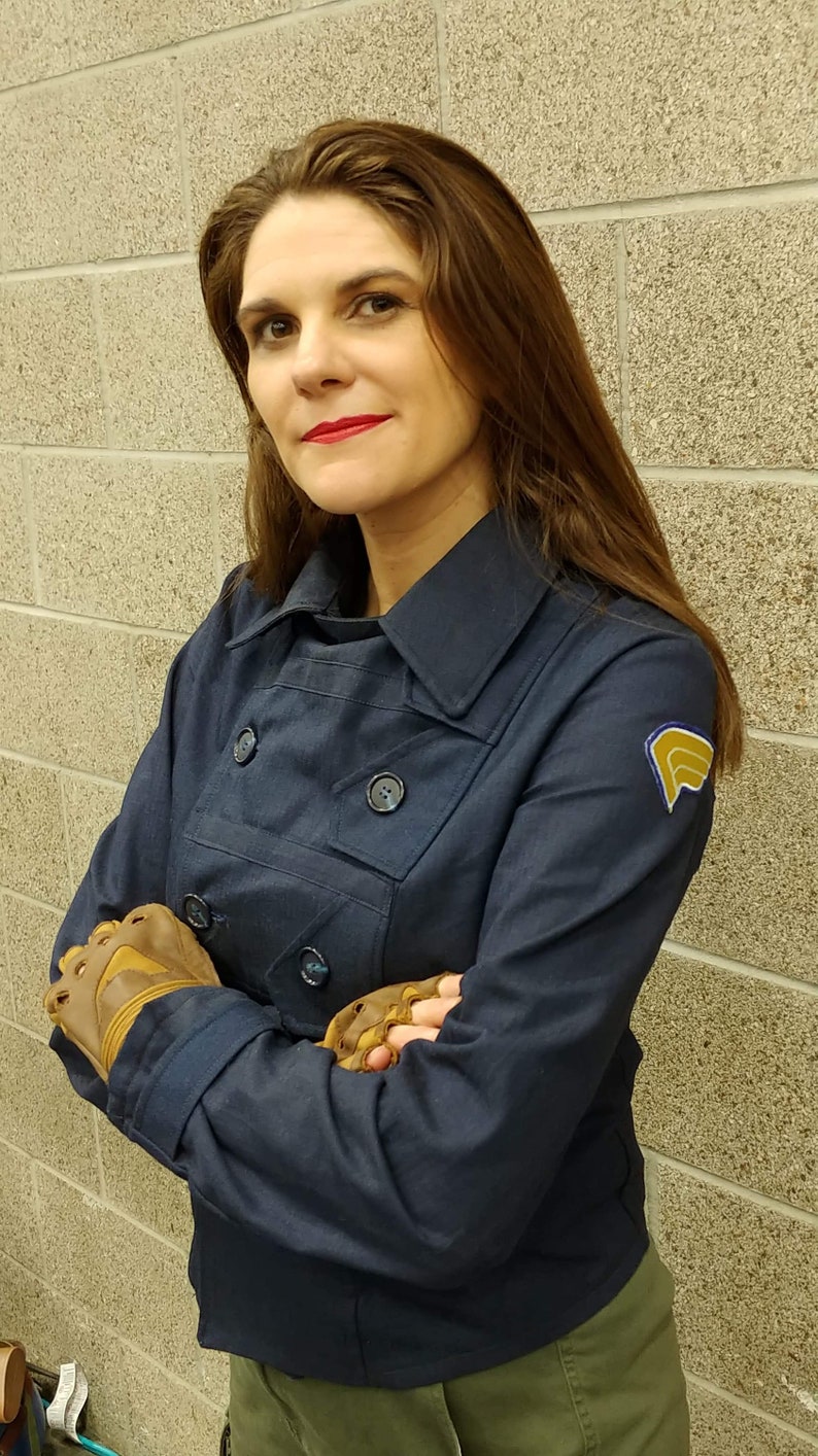 May include: A dark navy blue double-breasted jacket with a structured collar, long sleeves, and dark buttons. The jacket features a yellow and blue embroidered patch on the left shoulder. The garment is paired with brown fingerless leather gloves.