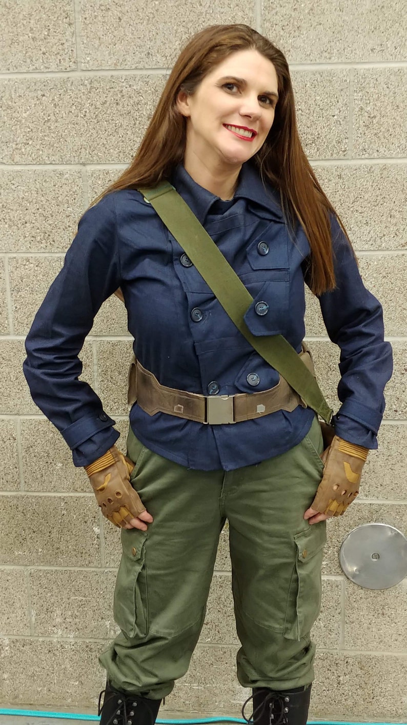 May include: A navy blue double-breasted jacket with a collar and buttons, paired with olive green cargo pants featuring side pockets. A wide brown utility belt with a metallic buckle is worn at the waist, complemented by a green shoulder strap and tan fingerless gloves.