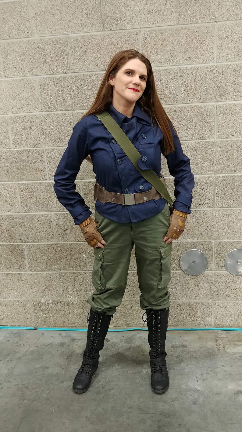 May include: A dark blue button-up jacket with a collar, olive green cargo pants with side pockets, a brown utility belt with a silver buckle, a green shoulder strap, tan fingerless gloves, and black lace-up boots reaching mid-calf.