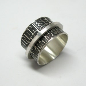 May include: A silver spinner ring with the words "KINDNESS" and "CONFIDENCE" etched in black. The ring has a rotating outer band and a polished finish, set against a white background.
