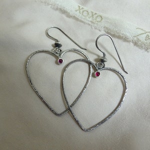 May include: A pair of silver heart-shaped earrings with small red gemstones in the center of each heart.