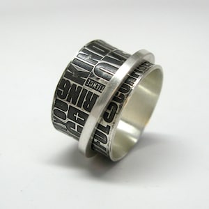 May include: A silver-colored ring with a textured band featuring the words "BEAUTIFUL", "RING", and other words in a block font. The ring has a spinning outer band and a polished silver finish.