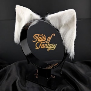 Może przedstawiać: Para białych, puszystych uszu kota przymocowanych do czarnej opaski na głowę. Opaska ma złoty napis "Tails of Fantasy". Uszy są wyprostowane i wyróżniają się na czarnym tle.