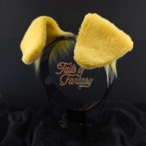 Pode incluir: Uma tiara preta com orelhas de coelho amarelas e fofas. A tiara tem o texto "Tails of Fantasy" em letras douradas. As orelhas são de um amarelo vibrante e são feitas de um material macio e felpudo.
