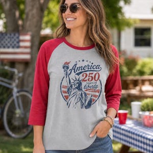 Puede incluir: Una camiseta estilo b&eacute;isbol gris jaspeado con mangas rojas de tres cuartos. El frente presenta un gr&aacute;fico azul de la Estatua de la Libertad con el texto America 250 years of liberty en un dise&ntilde;o circular con detalles en rojo y azul.