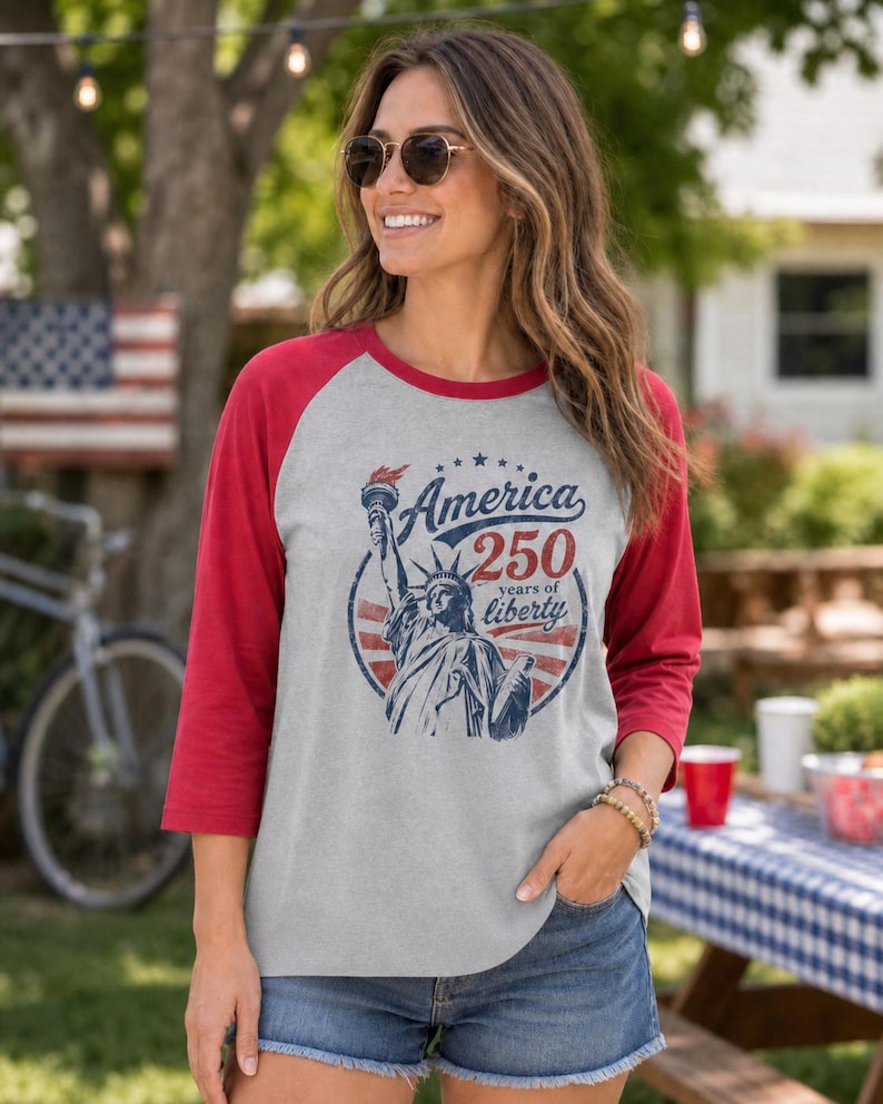 Puede incluir: Una camiseta estilo b&eacute;isbol gris jaspeado con mangas rojas de tres cuartos. El frente presenta un gr&aacute;fico azul de la Estatua de la Libertad con el texto America 250 years of liberty en un dise&ntilde;o circular con detalles en rojo y azul.