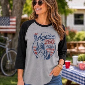 Puede incluir: Una camiseta gris de estilo b&eacute;isbol con mangas negras de tres cuartos. El frente presenta un gr&aacute;fico azul y rojo de la Estatua de la Libertad con el texto America 250 years of liberty dispuesto en un dise&ntilde;o circular con estrellas.
