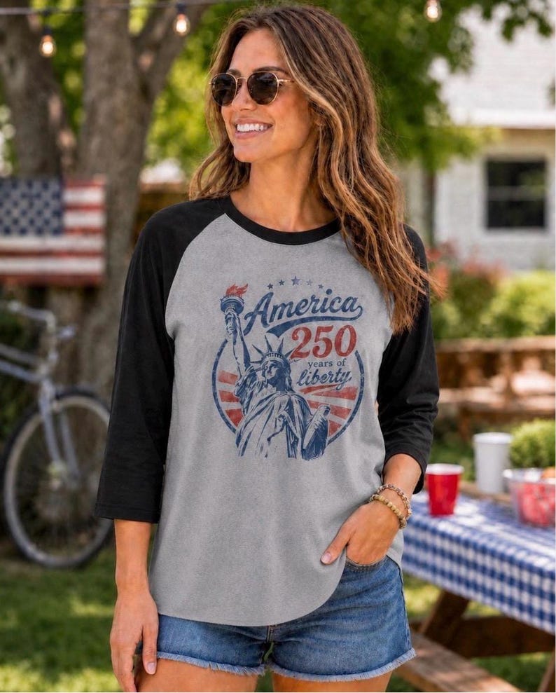 Puede incluir: Una camiseta gris de estilo b&eacute;isbol con mangas negras de tres cuartos. El frente presenta un gr&aacute;fico azul y rojo de la Estatua de la Libertad con el texto America 250 years of liberty dispuesto en un dise&ntilde;o circular con estrellas.