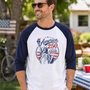 Puede incluir: Una camiseta estilo b&eacute;isbol blanca y azul marino con mangas tres cuartos. El frente presenta un gr&aacute;fico de la Estatua de la Libertad dentro de un marco circular con el texto America 250 years of liberty en tinta roja y azul.