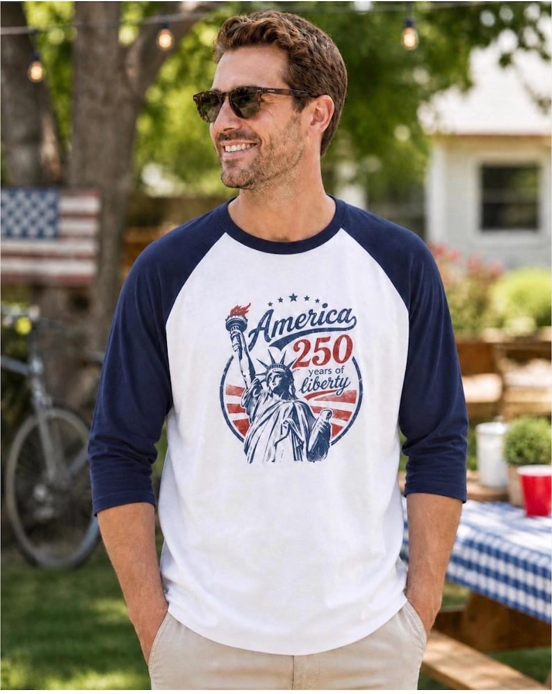 Puede incluir: Una camiseta estilo b&eacute;isbol blanca y azul marino con mangas tres cuartos. El frente presenta un gr&aacute;fico de la Estatua de la Libertad dentro de un marco circular con el texto America 250 years of liberty en tinta roja y azul.