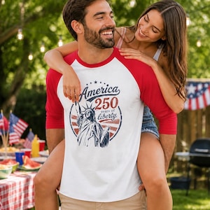 Puede incluir: Una camiseta blanca estilo b&eacute;isbol con mangas rojas de tres cuartos. El frente presenta un gr&aacute;fico de la Estatua de la Libertad dentro de un emblema circular con el texto America 250 years of liberty en tinta azul y roja.