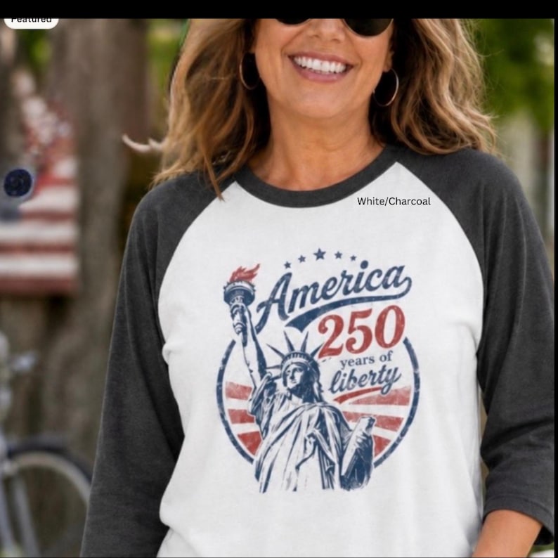 Puede incluir: Una camiseta de b&eacute;isbol ragl&aacute;n blanca y gris carb&oacute;n con un gr&aacute;fico de la Estatua de la Libertad en azul y rojo. El texto dice America 250 years of liberty. El dise&ntilde;o es circular con rayas rojas y estrellas azules celebrando el aniversario.