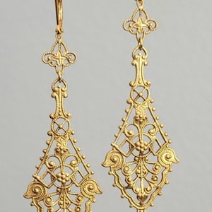 May include: A pair of ornate gold-colored earrings. Each earring features an intricate filigree design with floral and geometric patterns. The earrings have a long, teardrop shape, with a decorative top and a hook closure. The earrings are displayed against a neutral background.