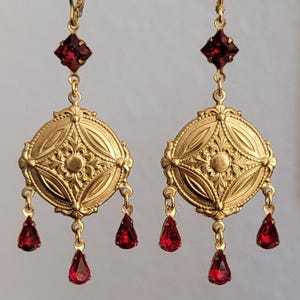 May include: A pair of gold-toned dangle earrings featuring a circular medallion with intricate embossed floral patterns. Each earring is accented with a square-cut red stone at the top and three dangling teardrop-shaped red stones at the base.
