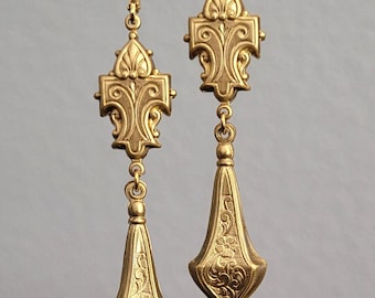 May include: A pair of gold-colored, ornate drop earrings. Each earring features a decorative top with a fleur-de-lis design, connected to a long, teardrop-shaped pendant with floral engravings. The earrings are suspended from a decorative bar.