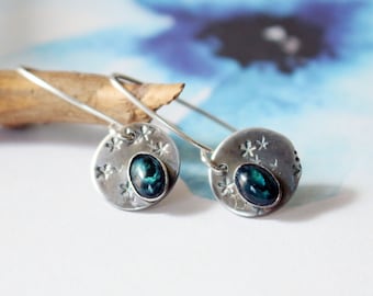 May include: Silver dangle earrings with a round, textured disc featuring a teal-colored gemstone and a snowflake design.
