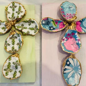 Könnte beinhalten: Zwei dekorative Kreuze aus Austernmuscheln. Das Kreuz links hat ein grünes und weißes Blumenmuster. Das Kreuz rechts hat ein blaues, rosa und weißes Blumenmuster.