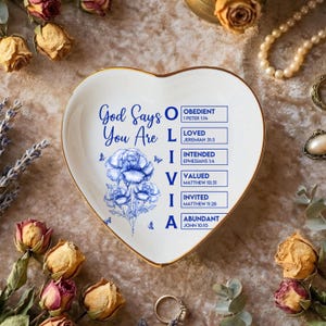 Puede incluir: Plato de cerámica en forma de corazón con borde dorado. El plato presenta obras de arte florales azules y el texto "Dios dice que eres AMADO, INTENCIONADO, VALORADO, INVITADO, ABUNDANTE." Elementos decorativos incluyen rosas secas y perlas.