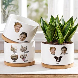 May include: White ceramic plant pots with custom faces of people and pets. The pots sit on wooden saucers. One pot contains a snake plant. The faces include Liam, Emma, Snowy, Marble, Max, Bella, and Milo.