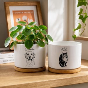 May include: Two white ceramic planters with wooden saucers. One planter features a dog illustration and the name "Charlie", the other has a cat illustration and the name "Milo". Both planters hold green plants.