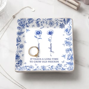 Puede incluir: Una bandeja cuadrada de cerámica blanca con un diseño floral azul. La bandeja presenta dos ilustraciones de flores azules, los nombres "Madison" y "Jackson", y la cita "IT TAKES A LONG TIME TO GROW OLD FRIENDS". Un anillo dorado descansa sobre la bandeja.