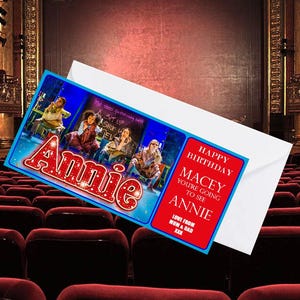 May include: A birthday card featuring the musical "Annie", with a blue border and the name in red, glittery letters. The card reads "Happy Birthday Macey, You're going to see Annie". It is presented inside a white envelope, in a theatre setting.