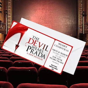 May include: A white envelope with a red and white card featuring the text "THE DEVIL WEARS PRADA A NEW MUSICAL". The card also includes "MERRY CHRISTMAS" and a personalized message. The background shows a theatre.