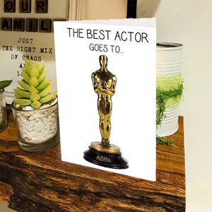 May include: A white card with the text "The Best Actor Goes To..." and a gold statuette of an Oscar award with the name "Alani" on the base.