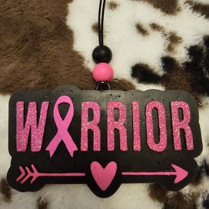 May include: A black rectangular hanging ornament with the word WARRIOR in pink glittery letters, where the letter A is replaced by a pink awareness ribbon. Below the text is a pink arrow pointing right with a pink heart in the center.