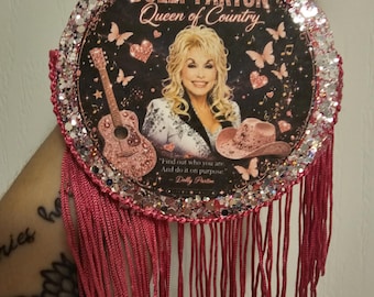 May include: A circular hanging ornament featuring a portrait of Dolly Parton with the text "DOLLY PARTON Queen of Country" and "Find out who you are. And do it on purpose. - Dolly Parton". The border is covered in pink glitter with pink fringe.
