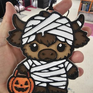 May include: A brown highland cow ornament wrapped in white bandages like a mummy, featuring large black eyes with white highlights and holding a glittery orange jack-o'-lantern pumpkin bucket, suspended by a thin black cord for hanging.