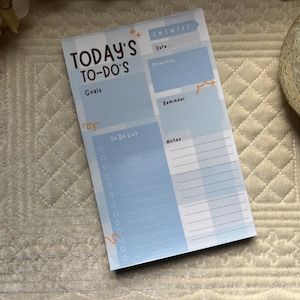 May include: A light blue and white notepad with the text "TODAY'S TO-DO'S" at the top. The notepad includes sections for goals, a to-do list, date, priorities, reminders, and notes. The notepad is rectangular and has a simple, clean design.