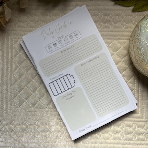 May include: A white daily check-in paper planner sheet with light green sections. It features a mood tracker with emojis, a section to write about feelings, an energy level battery icon, a space for needs, a pride section, and a daily goal line.