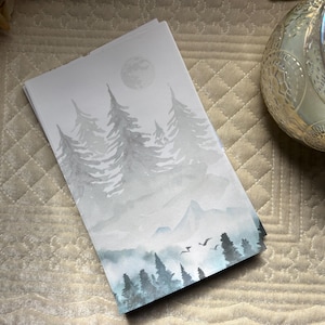 May include: A stack of notepads featuring a watercolor landscape with pine trees, mountains, and a full moon in shades of gray and blue. The artwork evokes a serene forest scene. The notepads are stacked on a textured, light-colored surface.