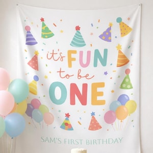 Op de afbeelding: Een witte verjaardagsbanner met kleurrijke feesthoeden en confetti. De banner leest "it's FUN to be ONE" en "SAM'S FIRST BIRTHDAY". Pastelkleurige ballonnen en een kleine taart zijn ook zichtbaar.