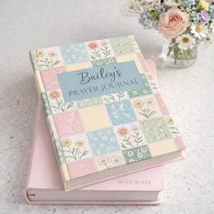 May include: A floral-patterned prayer journal with the text "Bailey's Prayer Journal" on the cover, resting atop a pink book labeled "Holy Bible." The journal features a pastel patchwork design with daisies and other flowers.