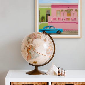 May include: A vintage globe sits on a white shelf with two wicker baskets. A framed photograph of colorful houses with a blue car in front of them is on the wall behind the globe.