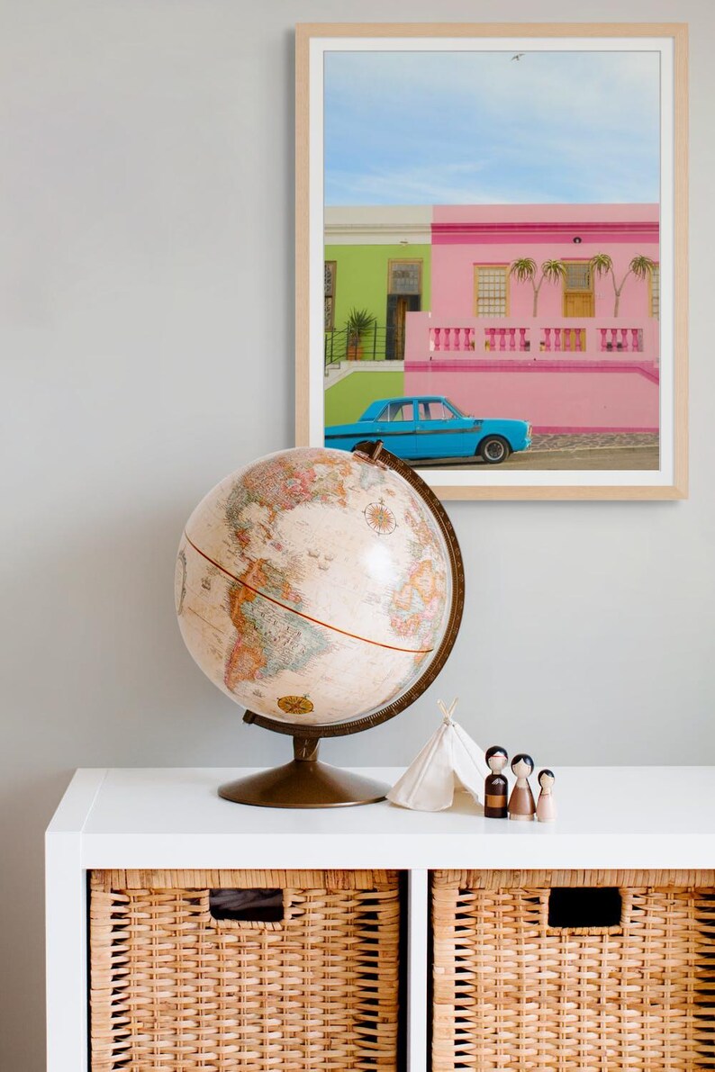 May include: A vintage globe sits on a white shelf with two wicker baskets. A framed photograph of colorful houses with a blue car in front of them is on the wall behind the globe.