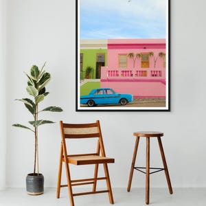 May include: A blue car parked in front of a colorful house with pink and green walls. The house has a white railing and palm trees in front of it. The sky is blue with a few white clouds.