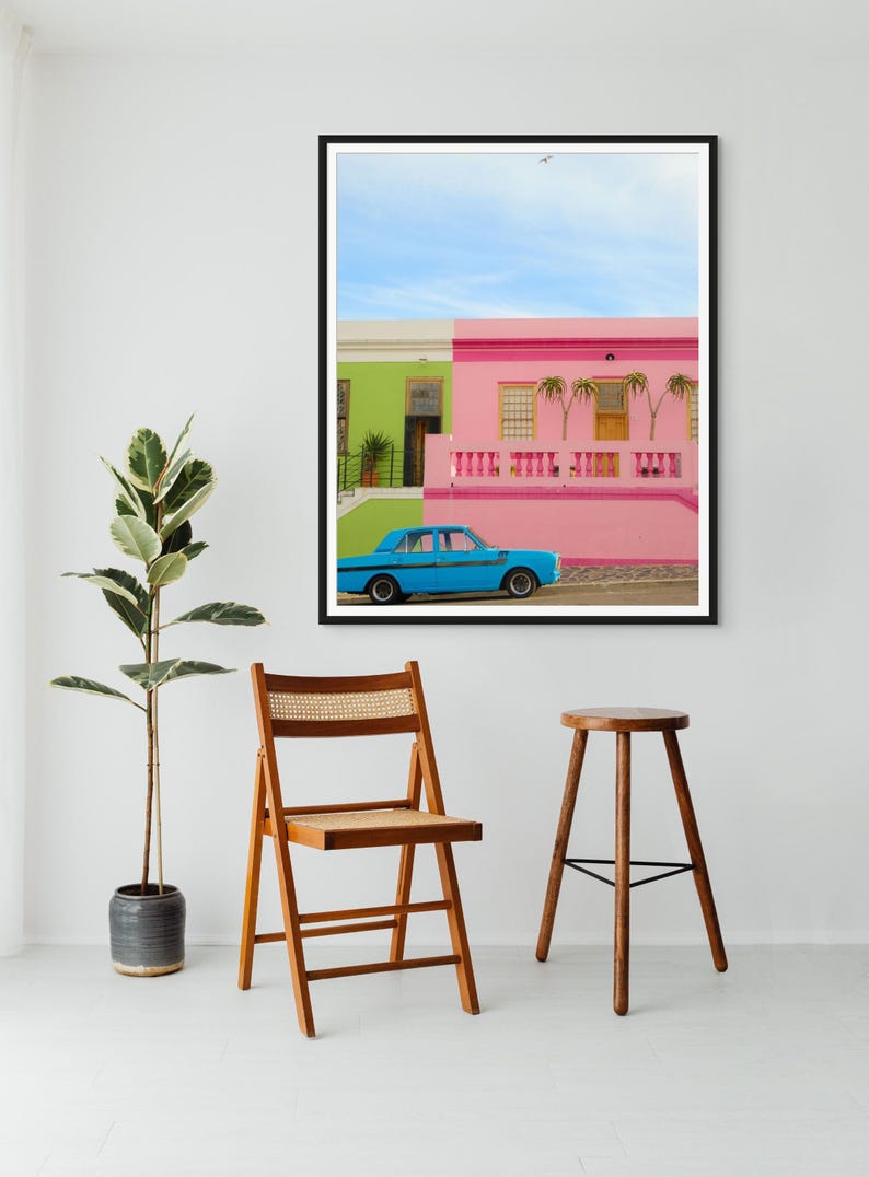 May include: A blue car parked in front of a colorful house with pink and green walls. The house has a white railing and palm trees in front of it. The sky is blue with a few white clouds.