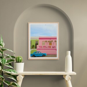 May include: A blue car parked in front of a pink and green building with palm trees. The building has a white railing and a white door.