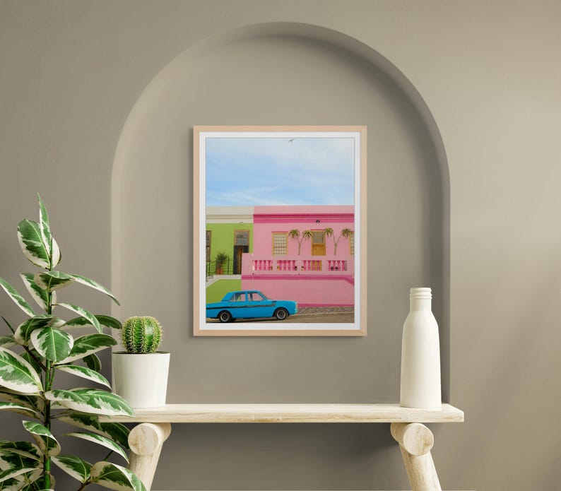 May include: A blue car parked in front of a pink and green building with palm trees. The building has a white railing and a white door.
