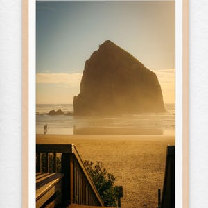 May include: A wooden staircase leads down to a sandy beach with a large rock formation in the distance. The sky is a soft orange and yellow, and the water is calm and clear.