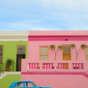 May include: A bright blue vintage car parked in front of a pink and green house with a white trim. The house has a balcony with a pink railing and two palm trees.