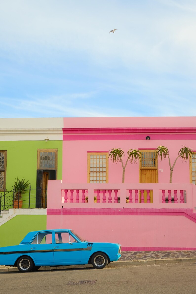 May include: A bright blue vintage car parked in front of a pink and green house with a white trim. The house has a balcony with a pink railing and two palm trees.