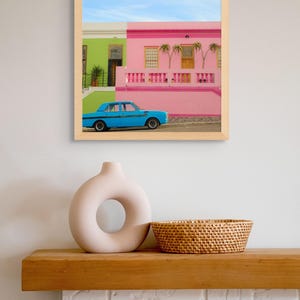 May include: A framed photograph of a blue car parked in front of a pink and green house. The house has a white railing and a palm tree in front of it. The sky is blue and there are clouds in the background.