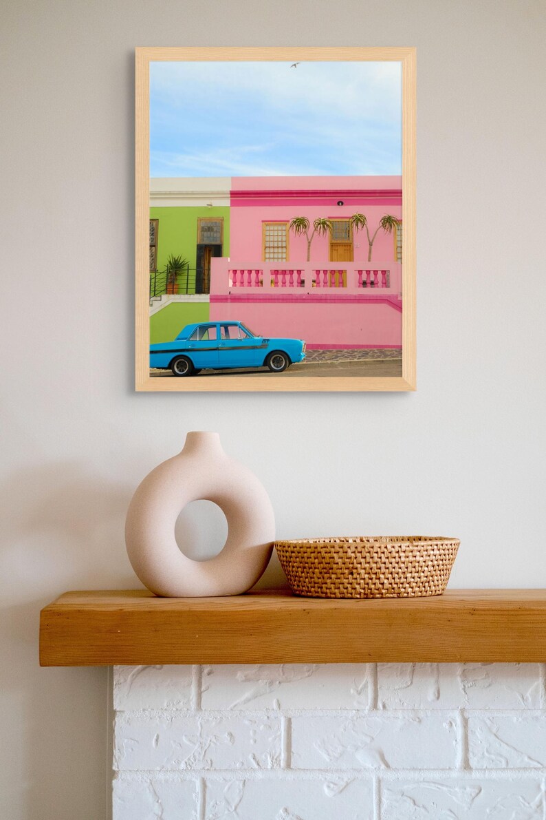 May include: A framed photograph of a blue car parked in front of a pink and green house. The house has a white railing and a palm tree in front of it. The sky is blue and there are clouds in the background.