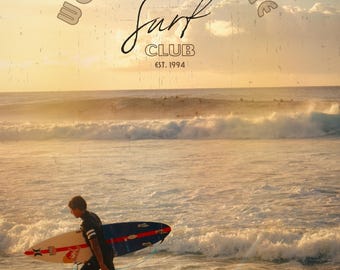 May include: A surfer walks along a sandy beach carrying a surfboard with a red bull logo. The ocean is in the background with waves crashing on the shore. The sky is a vibrant orange and yellow, with a vintage-style graphic overlay that reads "Welcome to the Surf Club Est. 1994". The text "Solivagant Photograph" is at the bottom of the image.
