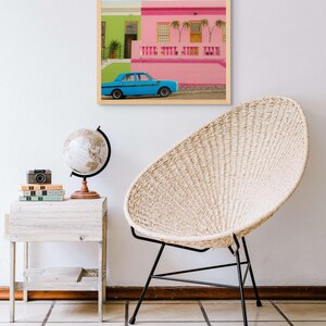 May include: A framed photograph of a blue car parked in front of a pink and green building. The building has a white railing and palm trees in front of it.