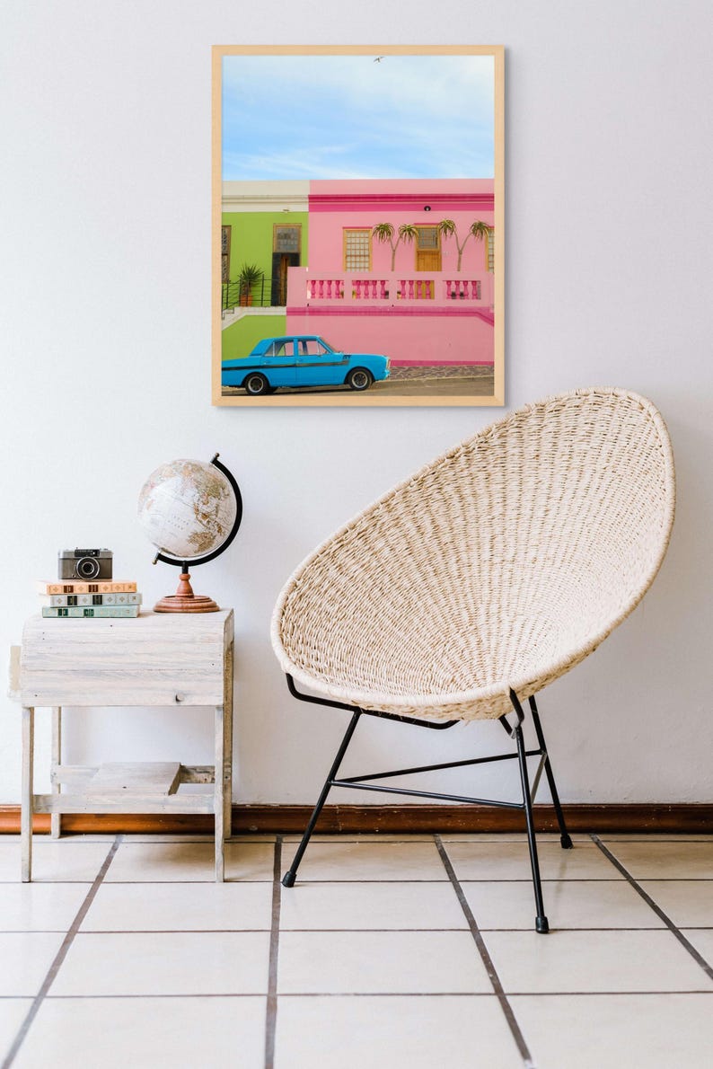 May include: A framed photograph of a blue car parked in front of a pink and green building. The building has a white railing and palm trees in front of it.